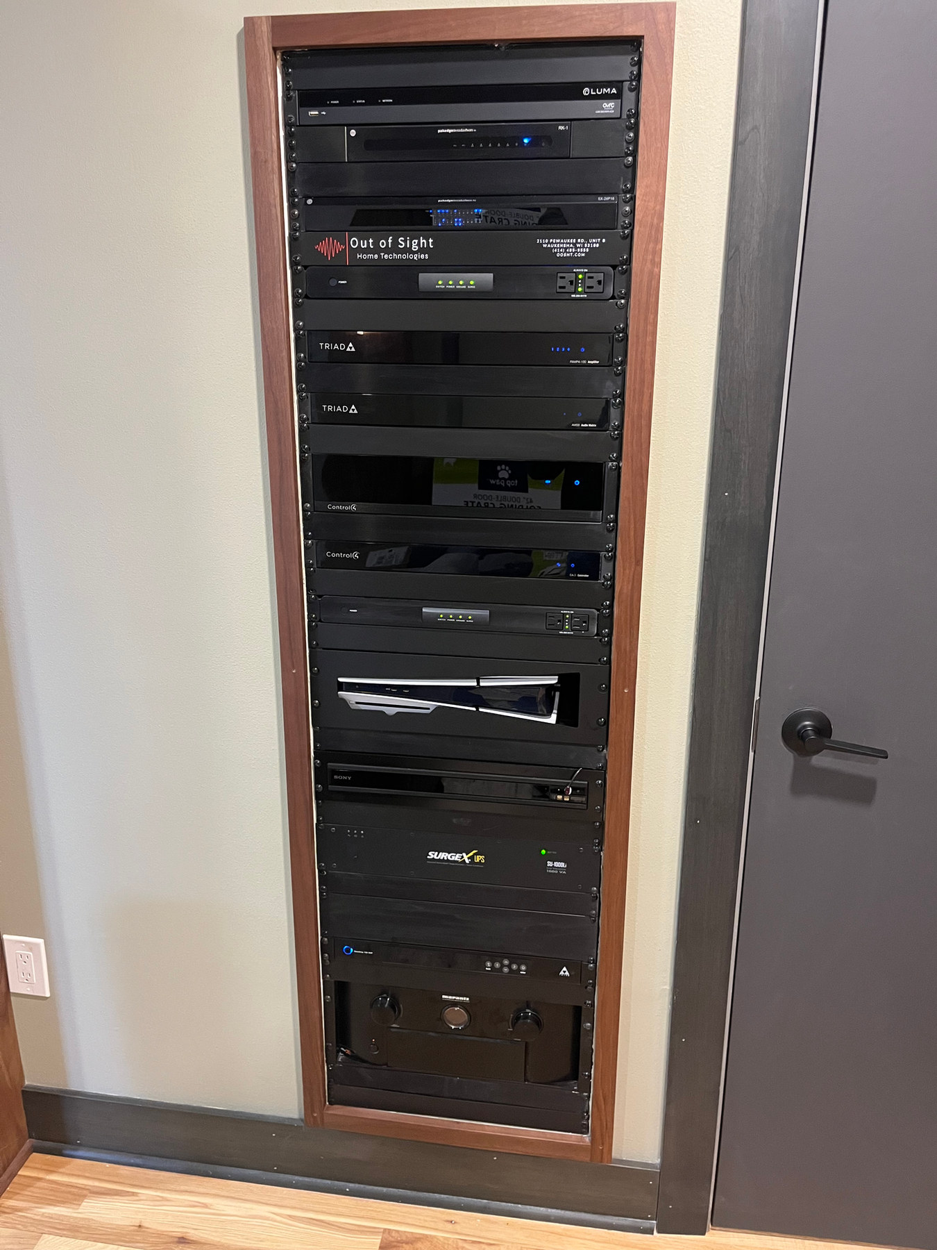 An equipment rack recessed into a walnut frame in the wall, carrying Control4, Triad, and Luma gear behind a labeled plate.